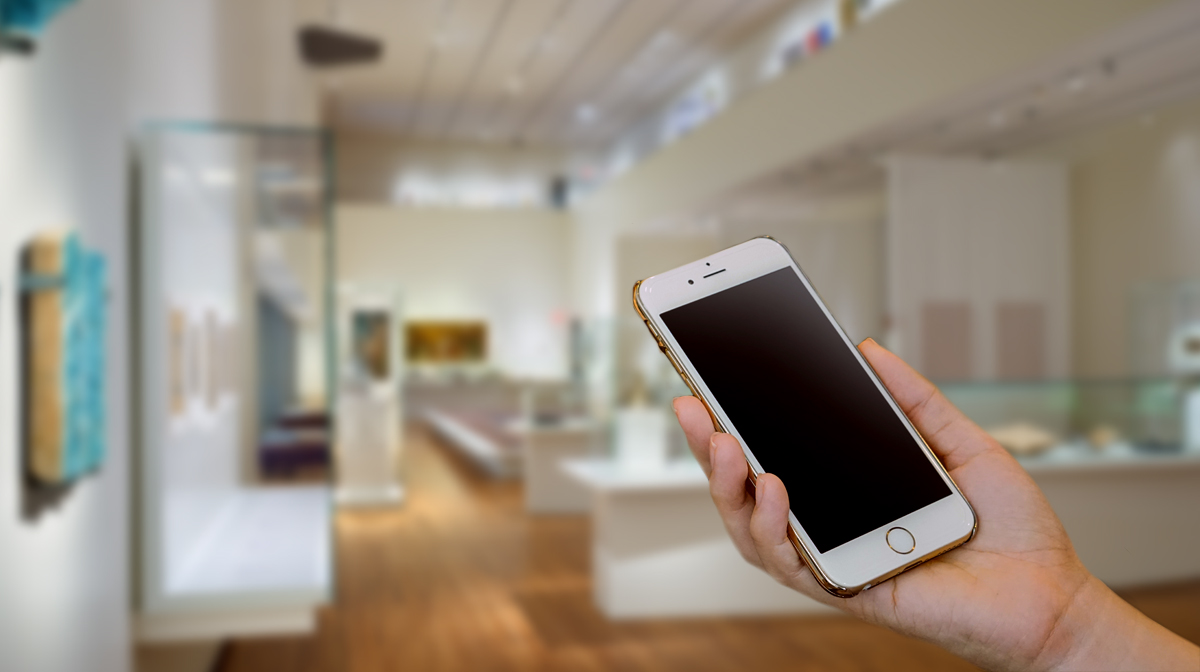 A hand holding a cell phone inside a museum gallery with glass cases and centuries-old artifacts from around the Islamic world. 